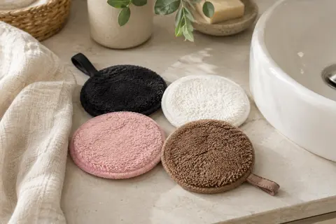 Quatre disques démaquillants ronds en microfibre, de couleurs variées, posés près d’un lavabo.