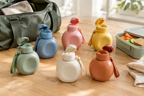 Six petites gourdes pour enfants aux coloris variés, avec bouchon à rabat et sangle, posées près d’un sac.